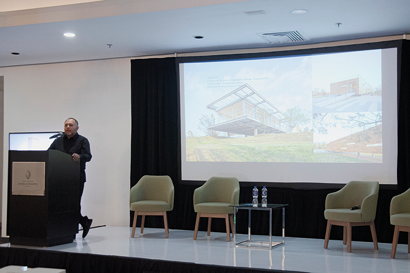 Un conferenciante especializado se presenta ante un selecto auditorio en un moderno salón de conferencias. En el escenario, el mobiliario minimalista -tres sillas exclusivas y una mesa con agua premium- subraya la atención al detalle y la calidad del entorno. La gran pantalla central proyecta información e imágenes sobre "Arquitectura del Nuevo Siglo", destacando innovaciones en diseño, sostenibilidad y lujo. Esta presentación está dirigida a inversores y compradores exigentes que buscan oportunidades únicas en desarrollos inmobiliarios de alto nivel, donde la excelencia arquitectónica se fusiona con el estilo de vida sofisticado.