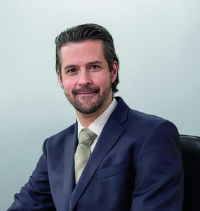 Un hombre de porte distinguido, con cabello oscuro y barba bien cuidada, luce un traje azul marino acompañado de camisa blanca y corbata a rayas. Sentado con serenidad ante un fondo claro y elegante, proyecta confianza y profesionalismo, el escenario ideal para abordar en profundidad las obligaciones clave de los propietarios de inmuebles premium. Esta imagen transmite credibilidad y preparación para orientar a inversores exigentes en la gestión eficiente y responsable de sus activos inmobiliarios.