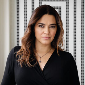 Una mujer de cabello castaño ondulado, vestida con un elegante top negro y un cuello distintivo, posa ante un fondo cómodo en blanco y negro. Su expresión serena refleja su interés por proyectos inmobiliarios frente al mar, espacios excepcionales que ofrecen vistas inigualables, privacidad y alto potencial de valorización. Esta imagen captura la esencia de quienes buscan inversiones exclusivas y residencias premium en ubicaciones privilegiadas.