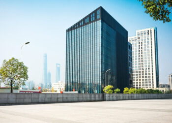 Un imponente edificio corporativo de diseño contemporáneo, con fachada de vidrio, se erige junto a una elegante torre blanca que evoca el estilo exclusivo de las residencias de marca en Baja. La plaza, perfectamente urbanizada y bordeada por árboles maduros, ofrece un ambiente sereno y cómodo. Al fondo, se perfilan rascacielos bajo un cielo despejado, completando un entorno urbano ideal para inversores o compradores que buscan propiedades premium en ubicaciones emblemáticas con proyección de alta plusvalía.