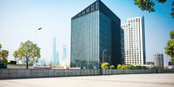 Un imponente edificio corporativo de diseño contemporáneo, con fachada de vidrio, se erige junto a una elegante torre blanca que evoca el estilo exclusivo de las residencias de marca en Baja. La plaza, perfectamente urbanizada y bordeada por árboles maduros, ofrece un ambiente sereno y cómodo. Al fondo, se perfilan rascacielos bajo un cielo despejado, completando un entorno urbano ideal para inversores o compradores que buscan propiedades premium en ubicaciones emblemáticas con proyección de alta plusvalía.