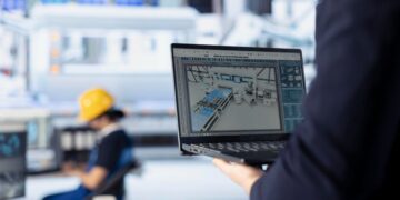Una persona sostiene un portátil que exhibe un modelo 3D de una instalación industrial de última generación, ilustrando el avance hacia la transformación digital. En segundo plano, difuminado, se observa a un operario con casco de seguridad interactuando con maquinaria especializada en un entorno industrial moderno. Esta escena resalta la integración de tecnología de punta en espacios industriales premium—una propuesta ideal para inversores y compradores visionarios que buscan activos inmobiliarios diseñados para maximizar la eficiencia operativa, la innovación y el valor sostenido.