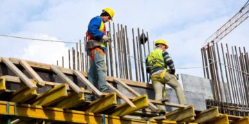 Dos trabajadores especializados, equipados con arneses de seguridad y cascos certificados, operan sobre la estructura de un edificio en desarrollo, donde destacan el acero expuesto de alta resistencia, vigas de madera seleccionada y un sistema de andamiaje profesional. Este entorno refleja una absoluta prioridad en protocolos avanzados de seguridad laboral, minimizando riesgos y garantizando la continuidad eficiente del proyecto. Estas prácticas aseguran estándares superiores en cada etapa constructiva, alineándose con las expectativas del segmento inmobiliario premium y proporcionando confianza a inversores y compradores que exigen calidad integral y máxima protección durante toda la ejecución.