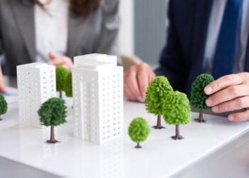 Dos ejecutivos, elegantemente vestidos, analizan en detalle una maqueta arquitectónica que destaca modernos edificios blancos rodeados de áreas verdes. En este entorno profesional, evalúan un proyecto inmobiliario de alto nivel enfocado en el diseño sostenible y el urbanismo responsable. Esta iniciativa no solo maximiza el valor patrimonial y la exclusividad para inversionistas y compradores exigentes, sino que también incorpora prácticas alineadas con la responsabilidad social y ambiental que definen las tendencias premium del sector.