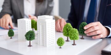 Dos ejecutivos, elegantemente vestidos, analizan en detalle una maqueta arquitectónica que destaca modernos edificios blancos rodeados de áreas verdes. En este entorno profesional, evalúan un proyecto inmobiliario de alto nivel enfocado en el diseño sostenible y el urbanismo responsable. Esta iniciativa no solo maximiza el valor patrimonial y la exclusividad para inversionistas y compradores exigentes, sino que también incorpora prácticas alineadas con la responsabilidad social y ambiental que definen las tendencias premium del sector.