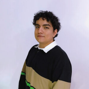 Un joven profesional, de imagen actual y distinguida—ataviado con un suéter de diseño contemporáneo en tonos negro, beige y verde sobre camisa blanca—posiciona su presencia ante un fondo neutro. Su actitud denota conciencia ambiental, cualidad esencial para quienes valoran inversiones inmobiliarias sostenibles y alineadas con las tendencias globales del mercado premium.