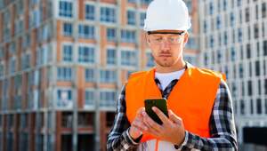 Un profesional de la construcción, equipado con casco blanco, gafas de seguridad y chaleco naranja, supervisa el avance de un exclusivo desarrollo inmobiliario a través de su teléfono inteligente. En el fondo, la estructura de un edificio premium en obra evidencia cómo la transformación digital está redefiniendo los estándares del sector inmobiliario en México. Esta integración tecnológica garantiza mayor eficiencia, precisión y transparencia en cada etapa del proceso constructivo, atributos claves para inversores y compradores que buscan proyectos residenciales o corporativos de alto valor patrimonial.