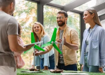 Un grupo de cuatro adultos se reúne en torno a una mesa que exhibe maquetas de aerogeneradores, en un entorno interior luminoso y moderno. Mientras uno de los asistentes sostiene documentación relevante, los demás interactúan y analizan activamente el potencial de las energías renovables como valor añadido para desarrollos inmobiliarios sostenibles. El espacio cuenta con amplios ventanales que permiten la entrada de luz natural y ofrecen vistas despejadas hacia áreas arboladas, aportando una atmósfera exclusiva que integra innovación, responsabilidad ambiental y máxima calidad en la experiencia residencial o inversora.