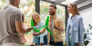 Un grupo de cuatro adultos se reúne en torno a una mesa que exhibe maquetas de aerogeneradores, en un entorno interior luminoso y moderno. Mientras uno de los asistentes sostiene documentación relevante, los demás interactúan y analizan activamente el potencial de las energías renovables como valor añadido para desarrollos inmobiliarios sostenibles. El espacio cuenta con amplios ventanales que permiten la entrada de luz natural y ofrecen vistas despejadas hacia áreas arboladas, aportando una atmósfera exclusiva que integra innovación, responsabilidad ambiental y máxima calidad en la experiencia residencial o inversora.