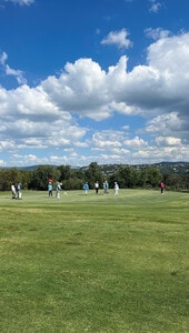 Un selecto grupo de personas se encuentra sobre el green impecablemente cuidado de un exclusivo campo de golf, enmarcado por suaves colinas y majestuosos árboles bajo un cielo parcialmente despejado. La calidad superior del césped resalta la excelencia del golf en México, consolidando su prestigio internacional como destino preferente para el turismo y la inversión inmobiliaria premium vinculada a campos de golf de alto nivel.