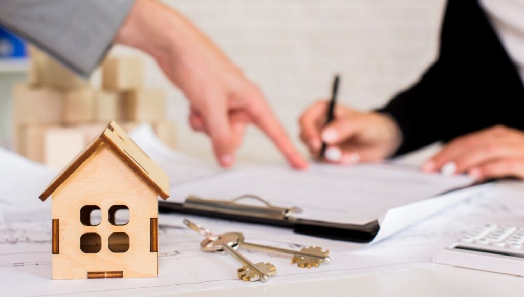 Sobre una mesa, un modelo arquitectónico en madera y llaves se disponen junto a documentos oficiales. Al fondo, dos personas revisan detenidamente los términos contractuales, enfatizando la formalidad y confianza del proceso de inversión inmobiliaria. Esta escena refleja el dinamismo actual del mercado premium de vivienda en renta en México, donde cada detalle es gestionado con rigor profesional para garantizar seguridad jurídica y sólidas oportunidades de retorno para compradores e inversionistas exigentes.