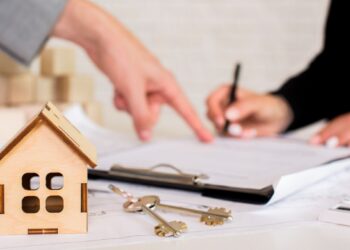 Sobre una mesa, un modelo arquitectónico en madera y llaves se disponen junto a documentos oficiales. Al fondo, dos personas revisan detenidamente los términos contractuales, enfatizando la formalidad y confianza del proceso de inversión inmobiliaria. Esta escena refleja el dinamismo actual del mercado premium de vivienda en renta en México, donde cada detalle es gestionado con rigor profesional para garantizar seguridad jurídica y sólidas oportunidades de retorno para compradores e inversionistas exigentes.