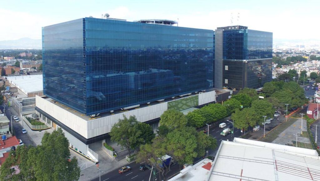 Vista aérea de un edificio corporativo de gran escala y arquitectura contemporánea, caracterizado por sus ventanales de cristal azul y operado por Fibra Uno. Rodeado de zonas arboladas y vialidades urbanas estratégicas, este inmueble integra diseño innovador con los más altos estándares en sostenibilidad, alineándose con los objetivos ESG hacia 2030. Un activo excepcional dentro del sector inmobiliario premium, ideal para inversionistas que buscan rentabilidad sólida y compromiso medioambiental en ubicaciones metropolitanas de alto crecimiento.