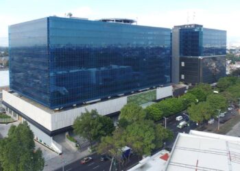Vista aérea de un edificio corporativo de gran escala y arquitectura contemporánea, caracterizado por sus ventanales de cristal azul y operado por Fibra Uno. Rodeado de zonas arboladas y vialidades urbanas estratégicas, este inmueble integra diseño innovador con los más altos estándares en sostenibilidad, alineándose con los objetivos ESG hacia 2030. Un activo excepcional dentro del sector inmobiliario premium, ideal para inversionistas que buscan rentabilidad sólida y compromiso medioambiental en ubicaciones metropolitanas de alto crecimiento.