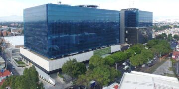Vista aérea de un edificio corporativo de gran escala y arquitectura contemporánea, caracterizado por sus ventanales de cristal azul y operado por Fibra Uno. Rodeado de zonas arboladas y vialidades urbanas estratégicas, este inmueble integra diseño innovador con los más altos estándares en sostenibilidad, alineándose con los objetivos ESG hacia 2030. Un activo excepcional dentro del sector inmobiliario premium, ideal para inversionistas que buscan rentabilidad sólida y compromiso medioambiental en ubicaciones metropolitanas de alto crecimiento.