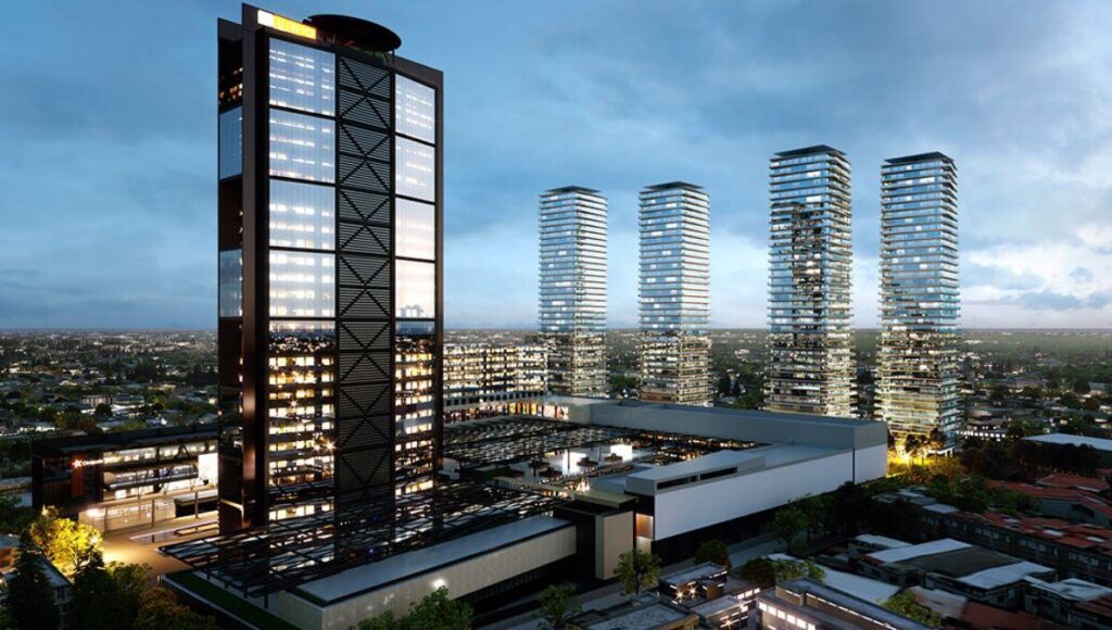 Un dinámico horizonte urbano al anochecer destaca una imponente torre de oficinas, cuyas ventanas iluminadas revelan la actividad empresarial de alto nivel, en conjunto con cuatro elegantes residencias verticales de cristal. Estas edificaciones premium reflejan la consolidación y el crecimiento sostenido de la representación inmobiliaria en mercados exclusivos. El entorno se complementa con edificaciones menores y un cuidadoso trazado vial, subrayando el desarrollo estratégico y moderno del sector para inversores y compradores que buscan ubicaciones privilegiadas, arquitectura vanguardista y alta plusvalía.
