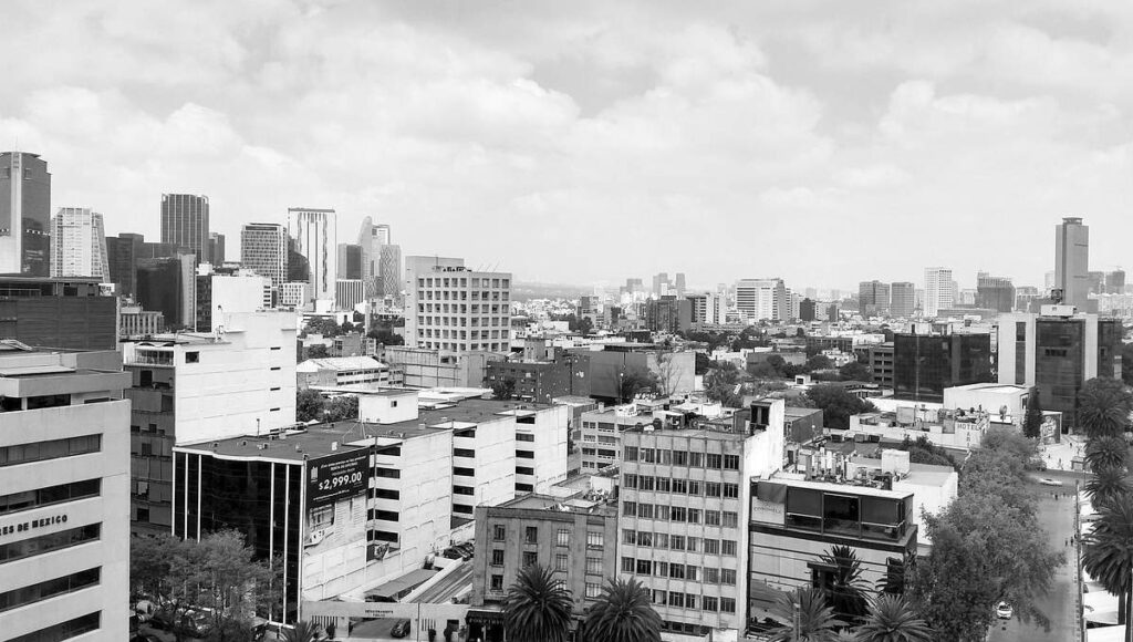 Fotografía en blanco y negro de un paisaje urbano dominado por imponentes rascacielos y elegantes edificios de media altura, reflejando el proceso de transformación que define las principales ciudades de México. La imagen captura un entorno metropolitano denso y cómodo, bajo un cielo nublado, donde la modernización continua ofrece oportunidades exclusivas para inversionistas exigentes que buscan participar en el desarrollo inmobiliario premium.