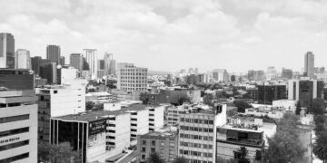 Fotografía en blanco y negro de un paisaje urbano dominado por imponentes rascacielos y elegantes edificios de media altura, reflejando el proceso de transformación que define las principales ciudades de México. La imagen captura un entorno metropolitano denso y cómodo, bajo un cielo nublado, donde la modernización continua ofrece oportunidades exclusivas para inversionistas exigentes que buscan participar en el desarrollo inmobiliario premium.