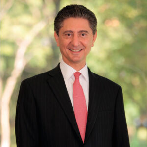 Un distinguido ejecutivo, ataviado con un impecable traje oscuro a rayas, camisa blanca y corbata roja, posa al aire libre delante de un entorno natural cuidadosamente difuminado. Su presencia transmite confianza y profesionalismo, representando a Fideicomiso de Turismo Los Cabos: una organización líder en el sector inmobiliario premium. Este portavoz simboliza el compromiso con la excelencia y la inversión segura en uno de los destinos más exclusivos para compradores e inversores exigentes que buscan propiedades de alto valor y rentabilidad asegurada.