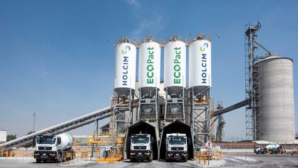 En una moderna instalación industrial dedicada a la producción de concreto de bajas emisiones, tres camiones mezcladores se encuentran alineados bajo imponentes silos identificados como Holcim México y Ecopact. Este entorno vanguardista incorpora tecnología de punta para el suministro eficiente y sostenible de materiales premium. La presencia de un silo principal y sofisticadas cintas transportadoras, enmarcadas por un cielo despejado, enfatiza la capacidad operativa e innovación del recinto: una garantía de calidad superior en soluciones constructivas para proyectos inmobiliarios exclusivos que exigen excelencia ambiental y estructural.