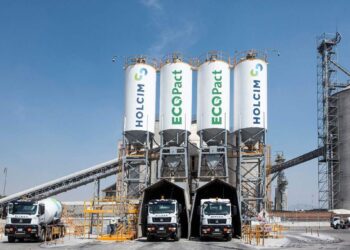 En una moderna instalación industrial dedicada a la producción de concreto de bajas emisiones, tres camiones mezcladores se encuentran alineados bajo imponentes silos identificados como Holcim México y Ecopact. Este entorno vanguardista incorpora tecnología de punta para el suministro eficiente y sostenible de materiales premium. La presencia de un silo principal y sofisticadas cintas transportadoras, enmarcadas por un cielo despejado, enfatiza la capacidad operativa e innovación del recinto: una garantía de calidad superior en soluciones constructivas para proyectos inmobiliarios exclusivos que exigen excelencia ambiental y estructural.