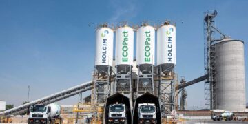 En una moderna instalación industrial dedicada a la producción de concreto de bajas emisiones, tres camiones mezcladores se encuentran alineados bajo imponentes silos identificados como Holcim México y Ecopact. Este entorno vanguardista incorpora tecnología de punta para el suministro eficiente y sostenible de materiales premium. La presencia de un silo principal y sofisticadas cintas transportadoras, enmarcadas por un cielo despejado, enfatiza la capacidad operativa e innovación del recinto: una garantía de calidad superior en soluciones constructivas para proyectos inmobiliarios exclusivos que exigen excelencia ambiental y estructural.