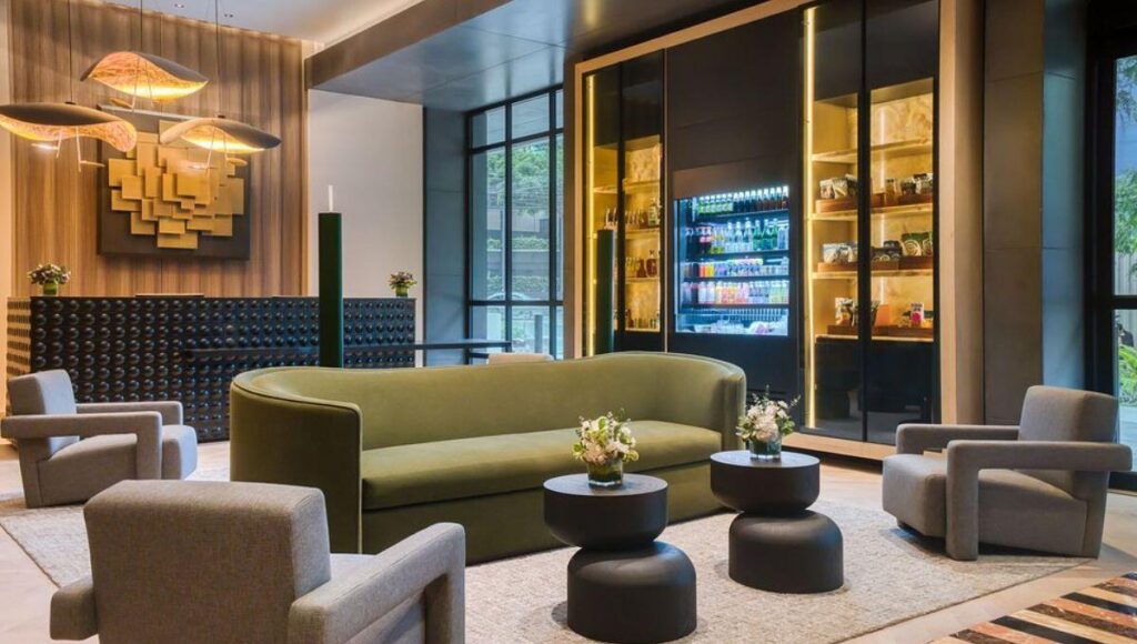 Lobby de hotel premium en Lima, Perú, diseñado para impresionar a los inversionistas y compradores más exigentes. Este espacio exclusivo integra un cómodo sofá curvo en tono verde, cuatro elegantes sillones grises y mesas redondas de diseño contemporáneo adornadas con arreglos florales selectos. La imponente obra de arte mural aporta un toque distintivo y memorable, mientras que la moderna vitrina refrigerada ofrece una selección gourmet de snacks para huéspedes distinguidos. Grandes ventanales bañan el ambiente con abundante luz natural, realzando los detalles arquitectónicos y aportando una atmósfera luminosa propia de los hoteles IHG más lujosos del sector inmobiliario premium en Lima.