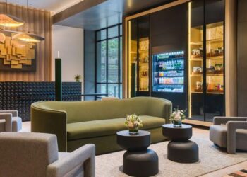 Lobby de hotel premium en Lima, Perú, diseñado para impresionar a los inversionistas y compradores más exigentes. Este espacio exclusivo integra un cómodo sofá curvo en tono verde, cuatro elegantes sillones grises y mesas redondas de diseño contemporáneo adornadas con arreglos florales selectos. La imponente obra de arte mural aporta un toque distintivo y memorable, mientras que la moderna vitrina refrigerada ofrece una selección gourmet de snacks para huéspedes distinguidos. Grandes ventanales bañan el ambiente con abundante luz natural, realzando los detalles arquitectónicos y aportando una atmósfera luminosa propia de los hoteles IHG más lujosos del sector inmobiliario premium en Lima.