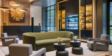 Lobby de hotel premium en Lima, Perú, diseñado para impresionar a los inversionistas y compradores más exigentes. Este espacio exclusivo integra un cómodo sofá curvo en tono verde, cuatro elegantes sillones grises y mesas redondas de diseño contemporáneo adornadas con arreglos florales selectos. La imponente obra de arte mural aporta un toque distintivo y memorable, mientras que la moderna vitrina refrigerada ofrece una selección gourmet de snacks para huéspedes distinguidos. Grandes ventanales bañan el ambiente con abundante luz natural, realzando los detalles arquitectónicos y aportando una atmósfera luminosa propia de los hoteles IHG más lujosos del sector inmobiliario premium en Lima.