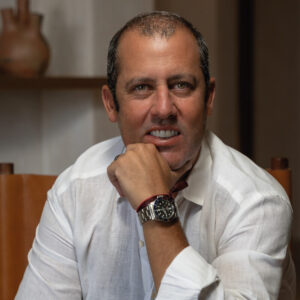 Un hombre de presencia distinguida, con barba prolija y cabello corto, viste una camisa blanca de alta calidad y un reloj elegante. En un entorno interior cuidadosamente diseñado, donde destacan detalles decorativos exclusivos, observa en actitud reflexiva. Su expresión evidencia el análisis profundo de los retos y las soluciones innovadoras que ofrece la arquitectura de vanguardia en Baja California: una región reconocida por su dinamismo inmobiliario premium y sus oportunidades únicas para inversionistas y compradores exigentes.