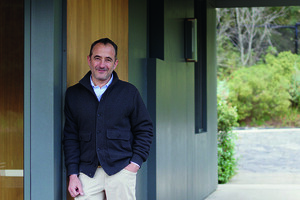 Un hombre vestido con un cárdigan oscuro y pantalones claros posa junto al marco de una puerta de madera, frente a un edificio moderno con certificación LEED. El entorno, rodeado de espacios verdes cuidadosamente integrados, refleja el compromiso con la sostenibilidad y el diseño cómodo que caracteriza a las propiedades premium más exclusivas del mercado inmobiliario. Este escenario resalta la fusión perfecta entre arquitectura contemporánea, eficiencia energética y un estilo de vida distinguido, ideal para inversores o compradores exigentes que priorizan calidad y responsabilidad ambiental en su próxima adquisición.