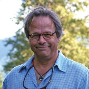 Hombre de mediana edad, cabello canoso y gafas, luce una camisa celeste a cuadros y un cuello. Se presenta en un entorno natural, rodeado de árboles y vegetación, transmitiendo serenidad y confianza a la cámara. Su amplia experiencia en Building Science respalda una visión innovadora para el desarrollo inmobiliario sostenible, orientada a inversores y compradores que valoran proyectos premium con estándares superiores de eficiencia, confort y respeto medioambiental.