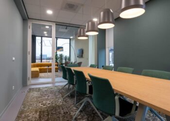 La sala de juntas, de diseño contemporáneo, destaca por su amplia mesa de madera, elegantes sillas verdes y luminarias colgantes que aportan sofisticación al espacio. Este ambiente ejemplifica la recuperación del sector inmobiliario corporativo premium en México rumbo a 2025. Las puertas de cristal dan paso a una exclusiva zona lounge con sofás amarillos y ventanas de gran formato, ofreciendo un entorno luminoso e inspirador que responde a las más altas expectativas de inversionistas y compradores exigentes.