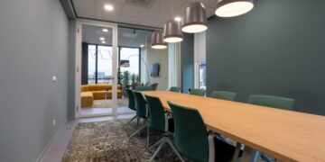 La sala de juntas, de diseño contemporáneo, destaca por su amplia mesa de madera, elegantes sillas verdes y luminarias colgantes que aportan sofisticación al espacio. Este ambiente ejemplifica la recuperación del sector inmobiliario corporativo premium en México rumbo a 2025. Las puertas de cristal dan paso a una exclusiva zona lounge con sofás amarillos y ventanas de gran formato, ofreciendo un entorno luminoso e inspirador que responde a las más altas expectativas de inversionistas y compradores exigentes.