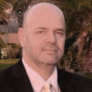 Un hombre de mediana edad, calvo y con barba perfectamente cuidada, viste un elegante traje y corbata mientras posa al aire libre con un entorno de exclusivas palmeras y exuberante vegetación de fondo. Representa al Fideicomiso de Turismo en Los Cabos, una organización clave en la promoción e impulso del sector inmobiliario premium en uno de los destinos más codiciados para inversionistas y compradores interesados.