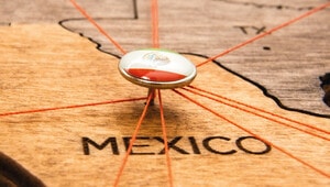 Un distintivo pin con la bandera de México señala estratégicamente una ubicación empresarial clave sobre un exclusivo mapa de madera. Desde este punto central irradian hilos naranjas que conectan con múltiples destinos, simbolizando la posición emergente de México como centro neurálgico para inversiones y operaciones premium, en el contexto global del nearshoring. Esta representación subraya las oportunidades únicas que ofrece el mercado inmobiliario mexicano para compradores e inversores exigentes: conectividad internacional, crecimiento sostenido y una plataforma sólida ante los nuevos retos económicos.