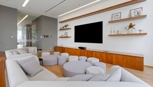 Sofisticada sala de estar en la Ciudad de México, diseñada para el segmento inmobiliario premium. El espacio integra elegantes sofás seccionales en tono gris claro, múltiples otomanas redondas y una pantalla plana de alta gama montada en la pared. Destacan estantes de madera con piezas decorativas seleccionadas y un mueble bajo que recorre toda la longitud del área audiovisual, brindando funcionalidad y estilo contemporáneo. Una opción ideal para inversionistas o compradores que buscan propiedades exclusivas dirigidas al mercado residencial de renta premium.