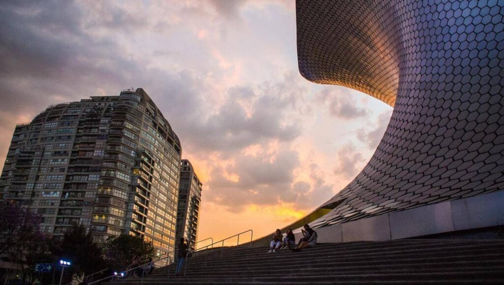 Un moderno rascacielos se alza junto a una estructura de elegante diseño curvilíneo y acabados en mosaico hexagonal, en el corazón de una de las colonias más exclusivas y codiciadas para vivir o invertir en la Ciudad de México. Bañada por la luz cálida del atardecer, esta escena refleja un entorno sofisticado donde residentes disfrutan espacios comunes integrados con arquitectura vanguardista y vistas privilegiadas; atributos que distinguen a las propiedades premium orientadas a compradores e inversionistas que buscan plusvalía, seguridad y calidad de vida incomparable.