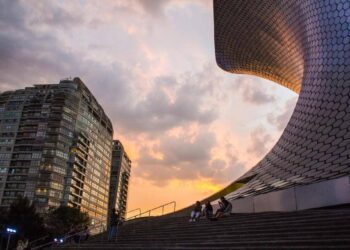 Un moderno rascacielos se alza junto a una estructura de elegante diseño curvilíneo y acabados en mosaico hexagonal, en el corazón de una de las colonias más exclusivas y codiciadas para vivir o invertir en la Ciudad de México. Bañada por la luz cálida del atardecer, esta escena refleja un entorno sofisticado donde residentes disfrutan espacios comunes integrados con arquitectura vanguardista y vistas privilegiadas; atributos que distinguen a las propiedades premium orientadas a compradores e inversionistas que buscan plusvalía, seguridad y calidad de vida incomparable.