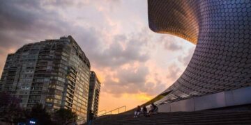 Un moderno rascacielos se alza junto a una estructura de elegante diseño curvilíneo y acabados en mosaico hexagonal, en el corazón de una de las colonias más exclusivas y codiciadas para vivir o invertir en la Ciudad de México. Bañada por la luz cálida del atardecer, esta escena refleja un entorno sofisticado donde residentes disfrutan espacios comunes integrados con arquitectura vanguardista y vistas privilegiadas; atributos que distinguen a las propiedades premium orientadas a compradores e inversionistas que buscan plusvalía, seguridad y calidad de vida incomparable.