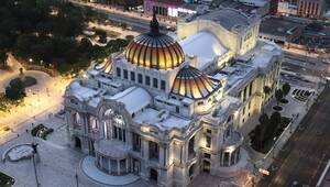 Vista aérea del Palacio de Bellas Artes, emblemático edificio de mármol blanco coronado por una cúpula en tonos naranja y dorado, enmarcado por áreas verdes y el vibrante entorno urbano al anochecer. Esta icónica referencia arquitectónica se ubica en proximidad inmediata a algunas de las colonias más exclusivas y cotizadas para invertir o residir en la Ciudad de México, integrando historia, cultura y plusvalía inigualable en el corazón de la capital.