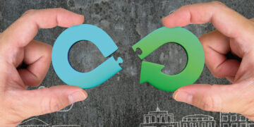 Dos manos sostienen piezas circulares de rompecabezas en azul y verde, diseñadas en forma de flechas que encajan perfectamente. El fondo oscuro, realzado por siluetas blancas de edificios, representa la innovadora iniciativa "Reciclar para Construir" de Saint-Gobain en Santa Catarina. Este proyecto integra sostenibilidad y vanguardia arquitectónica, ofreciendo desarrollos inmobiliarios premium que optimizan recursos y elevan los estándares medioambientales-una propuesta exclusiva para inversores y compradores que buscan excelencia, responsabilidad social y plusvalía en cada metro cuadrado.