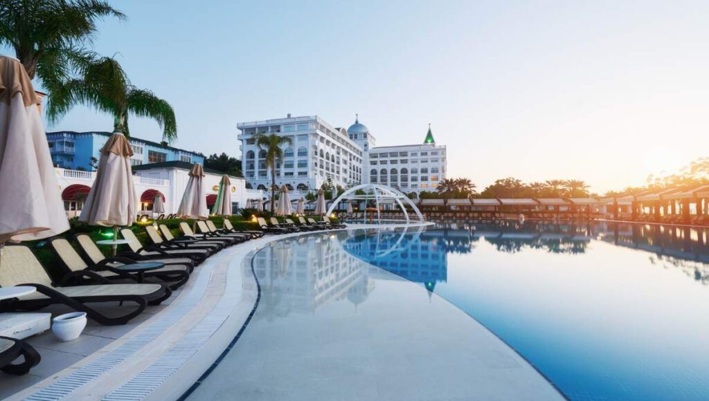 Una exclusiva piscina al aire libre, enmarcada por elegantes camastros y sombrillas cuidadosamente dispuestas, invita a disfrutar de un entorno de relajación y sofisticación. Al fondo, una imponente edificación hotelera de líneas blancas destaca entre majestuosas palmeras y cielos despejados al amanecer o atardecer. Esta propiedad forma parte del segmento premium del sector inmobiliario, ofreciendo un ambiente inigualable para inversores o compradores que valoran el confort, la privacidad y una experiencia única en destinos exclusivos.