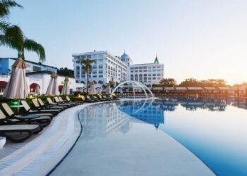 Una exclusiva piscina al aire libre, enmarcada por elegantes camastros y sombrillas cuidadosamente dispuestas, invita a disfrutar de un entorno de relajación y sofisticación. Al fondo, una imponente edificación hotelera de líneas blancas destaca entre majestuosas palmeras y cielos despejados al amanecer o atardecer. Esta propiedad forma parte del segmento premium del sector inmobiliario, ofreciendo un ambiente inigualable para inversores o compradores que valoran el confort, la privacidad y una experiencia única en destinos exclusivos.