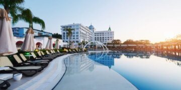 Una exclusiva piscina al aire libre, enmarcada por elegantes camastros y sombrillas cuidadosamente dispuestas, invita a disfrutar de un entorno de relajación y sofisticación. Al fondo, una imponente edificación hotelera de líneas blancas destaca entre majestuosas palmeras y cielos despejados al amanecer o atardecer. Esta propiedad forma parte del segmento premium del sector inmobiliario, ofreciendo un ambiente inigualable para inversores o compradores que valoran el confort, la privacidad y una experiencia única en destinos exclusivos.