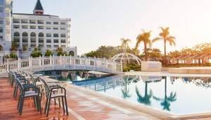 Zona exclusiva de alberca en hotel: elegantes camastros alineados sobre un deck de porcelanato, puente peatonal de diseño blanco y arcos que cruzan la piscina, rodeada de palmeras y con los edificios hoteleros como telón de fondo. Este entorno sofisticado no solo eleva el estándar visual y el atractivo para huéspedes premium, sino que es ideal para inversionistas que buscan activos diferenciados donde la integración de soluciones energéticas eficientes posibilita una significativa reducción en los costes operativos eléctricos, potenciando la rentabilidad y sostenibilidad del proyecto inmobiliario.