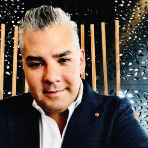 Un distinguido profesional del sector inmobiliario premium, de cabello canoso y porte elegante, viste un blazer oscuro y camisa blanca mientras reflexiona sobre los retos y las soluciones innovadoras en la arquitectura de la Baja. Sentado frente a una pared moderna cuyo diseño destaca por sus elegantes listones verticales de madera y atractivos paneles negros con patrones exclusivos, proyecta una visión estratégica orientada a satisfacer las expectativas de inversores y compradores que buscan propiedades excepcionales con alto valor patrimonial.