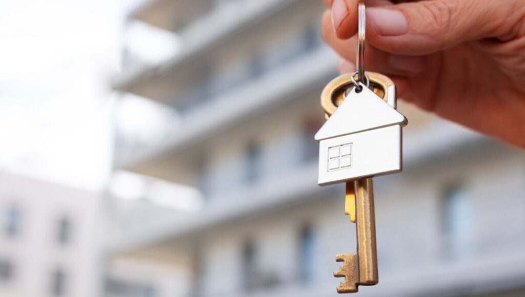 Una mano sostiene un llavero con un colgante en forma de casa y una llave, al frente de un exclusivo edificio residencial de fondo difuminado. Esta imagen simboliza el acceso a propiedades premium en desarrollos de usos mixtos modernos en Puebla, ofreciendo oportunidades excepcionales para inversores y compradores que buscan plusvalía, exclusividad y estilo de vida sofisticado.