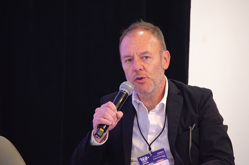 Un hombre elegantemente vestido con traje y camisa blanca, identificado por su distintivo de acceso, comparte su perspectiva sobre la innovación arquitectónica mientras interviene en un foro exclusivo del sector inmobiliario premium. Sentado en un entorno sofisticado y bajo una atmósfera monocromática que realza la gravedad de su discurso, transmite una visión transformadora para el desarrollo urbano. Su enfoque apunta a redefinir los estándares de las ciudades modernas, priorizando proyectos inmobiliarios vanguardistas que satisfacen las expectativas de inversores y compradores exigentes.