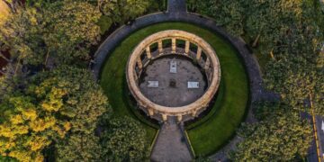 Vista aérea de un emblemático monumento circular de piedra, rodeado por jardines meticulosamente cuidados y frondosos árboles en el corazón del Centro Histórico de Guadalajara. Este espacio, recientemente renovado, se conecta armónicamente a través de elegantes senderos pavimentados que se dirigen al centro del monumento. Ubicación privilegiada y diseño arquitectónico icónico que realzan el valor patrimonial e inmobiliario, convirtiéndolo en un entorno ideal para inversiones exclusivas o proyectos residenciales premium.